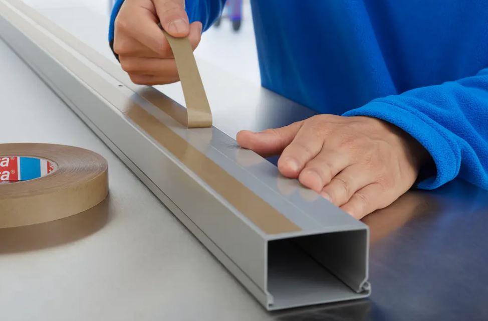 Jemand bringt präzise beigefarbenes Klebeband auf die Oberseite eines grauen U-Profils auf, das auf einer glänzenden Arbeitsfläche liegt. Links vorn liegt eine Rolle desselben Bands, bereit zur Nutzung.