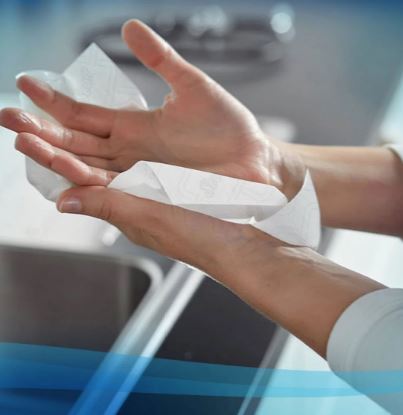Gros plan sur des mains utilisant une serviette en rouleau blanche gaufrée pour se sécher. Elles l'écartent pour absorber l'eau. À l'arrière-plan, on aperçoit un lavabo flou, ce qui souligne le caractère hygiénique de cette utilisation.