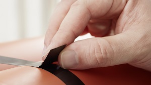 Eine rechte Hand bringt schwarzes Klebeband auf eine glänzende orange Oberfläche auf. Die Finger drücken das tesa Strapping-Klebeband präzise fest, um es auf einer abgerundeten Form zu fixieren. Dies zeigt manuelle Anwendung und starke Haftung.
