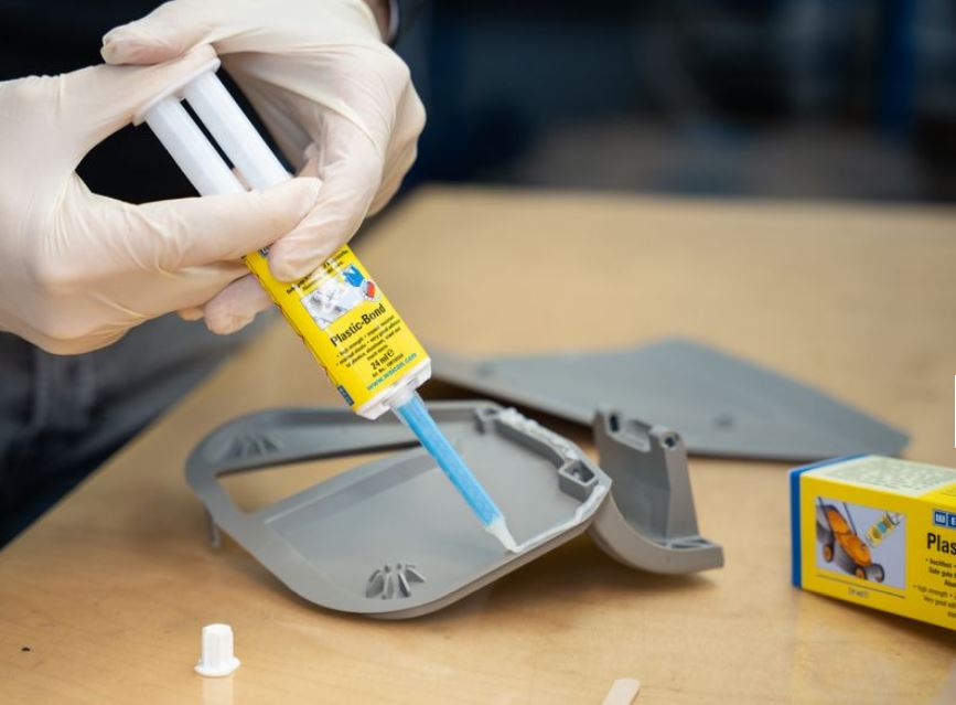 Eine behandschuhte Hand drückt einen gelben Strukturklebstoff-Spender mit blauer Spitze, der präzise eine weiße Klebstoffraupe auf die Kante eines grauen Kunststoffbauteils aufträgt. WEICON Plastic-Bond klebt Kunststoffteile.