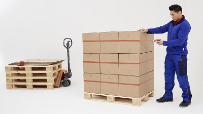 Ein Mann in blauer Arbeitskleidung sichert einen Stapel brauner Kartons auf einer Holzpalette mit orangefarbenem tesa Strapping-Klebeband. Drei waagerechte Bänder halten die Pakete stabil zusammen. Links steht eine leere Palette.