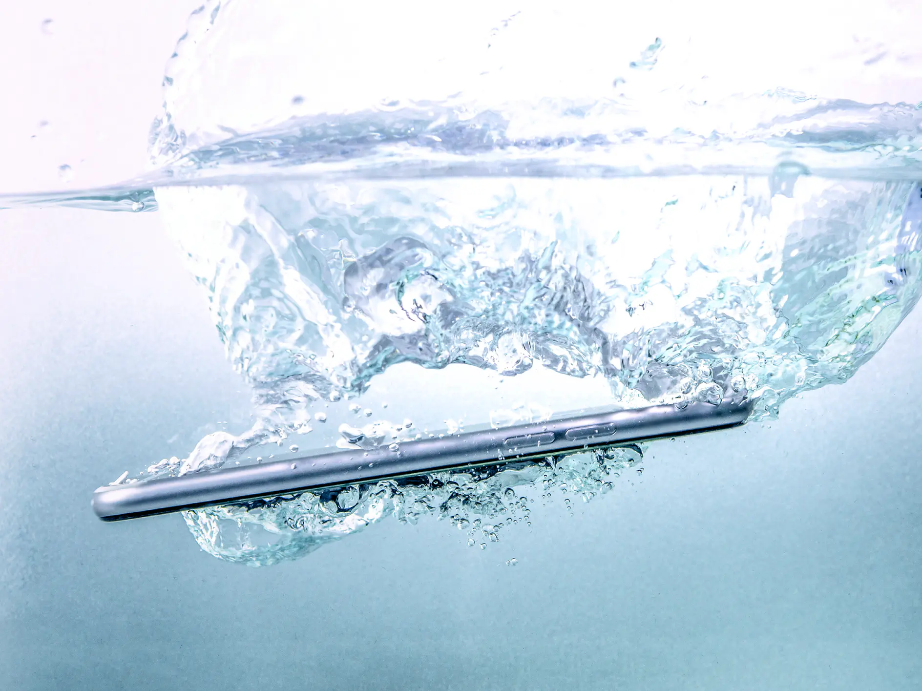 Un smartphone argenté plonge dans de l'eau claire, provoquant une grande vague et de nombreuses bulles d'air qui remontent à la surface. La moitié supérieure montre la surface, fortement agitée par l'impact.