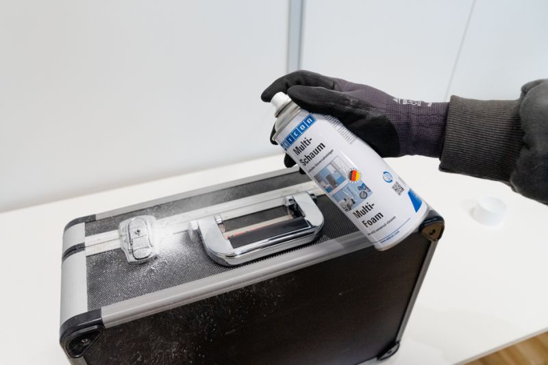 Eine Hand mit schwarzem Arbeitshandschuh sprüht weißen Reinigungsschaum aus einer WEICON Multi-Schaum Dose auf einen schwarzen Koffer mit silbernen Metallbeschlägen. Der dichte Schaum bedeckt bereits Teile der Oberfläche.