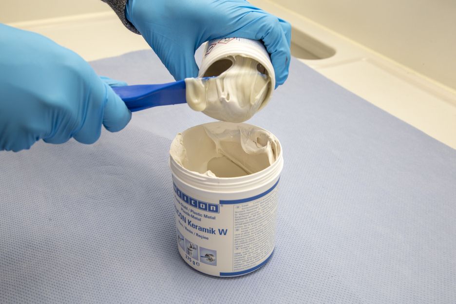 Blaue Handschuhe entnehmen weiße, pastöse Keramikmasse mit blauem Spachtel aus einem Eimer und füllen sie in einen anderen. Epoxidharz-System für Verschleißschutzbeschichtungen, cremige Konsistenz. "WEICON Keramik W".