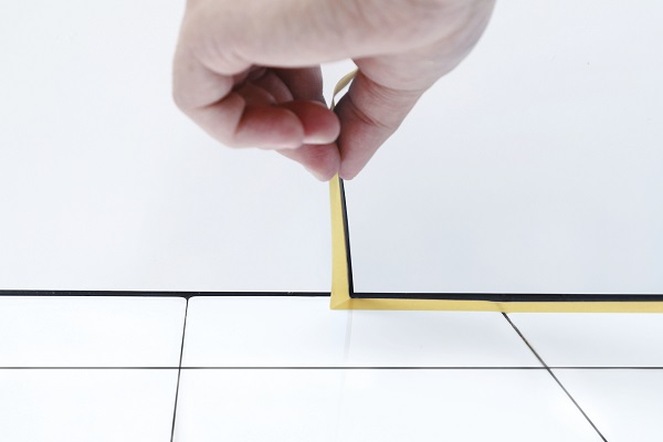 Eine Hand platziert ein schmales, goldfarbenes Abdeckband entlang der Kante zwischen weißer Wand und Bodenfliesen. Das 3M Super UV-Abdeckband sorgt für saubere Linien, präzises Abdecken innen/außen und lässt sich rückstandsfrei entfernen.