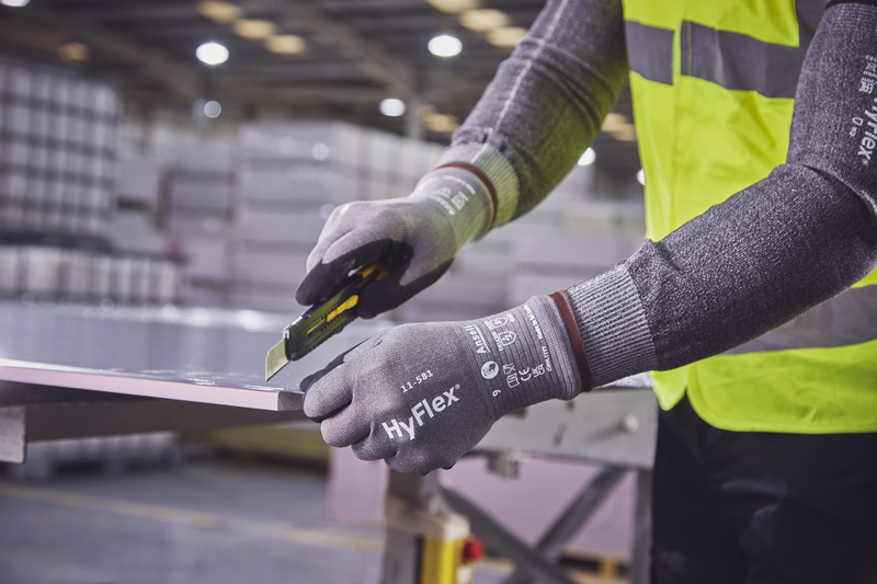 Nahaufnahme einer Person in grauen HyFlex Schnittschutzhandschuhen und gelber Warnweste, die mit einem Cuttermesser ein helles Plattenmaterial präzise schneidet. Hände geschützt, Klinge fährt an der Kante entlang. Lagerbereich im Hintergrund.