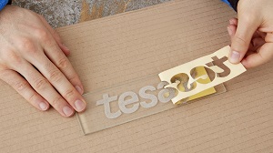 Zwei Hände arbeiten an einer Glasplatte, auf der „tesa“ sandgestrahlt/ geätzt sichtbar ist. Eine Hand zieht vorsichtig tesa 4434 Elefantenhaut-Abdeckband ab; der hellbraune Papierträger zeigt die ausgeschnittenen Buchstaben.