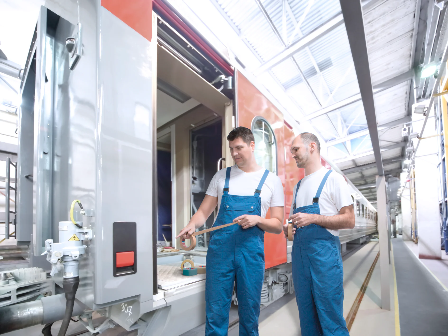 Deux techniciens en combinaisons bleues et t-shirts blancs sont dans un hangar de maintenance près d’un wagon ouvert. Celui de gauche tient du ruban adhésif et montre un détail, l’autre en tient aussi.