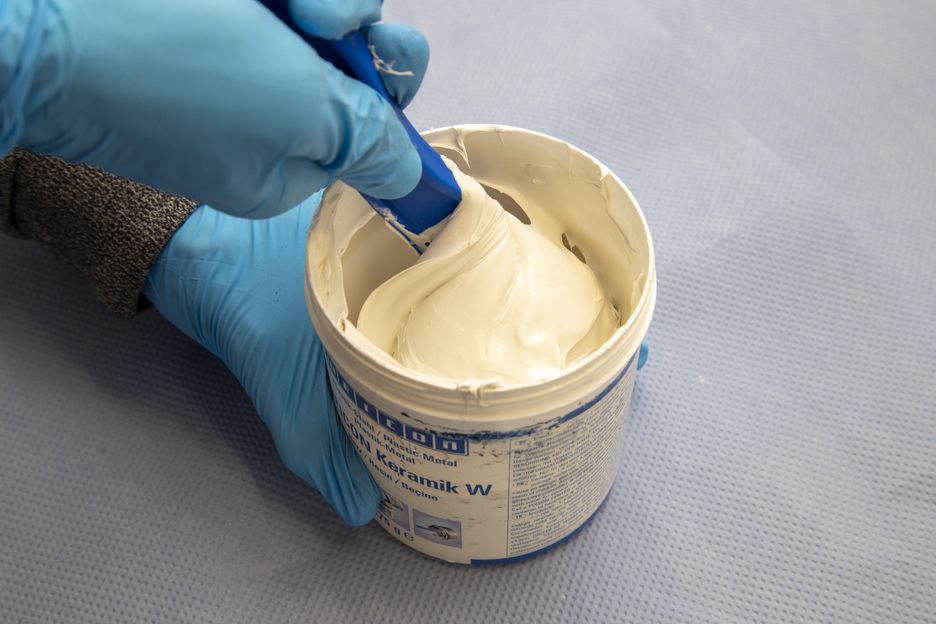 Eine Hand mit blauem Handschuh rührt mit blauem Spachtel in einem offenen weißen Becher mit cremiger, weißer WEICON Keramik W Epoxidharz-Masse. Die Paste ist spachtelbar und dick. Der Becher steht auf hellblauer Oberfläche.