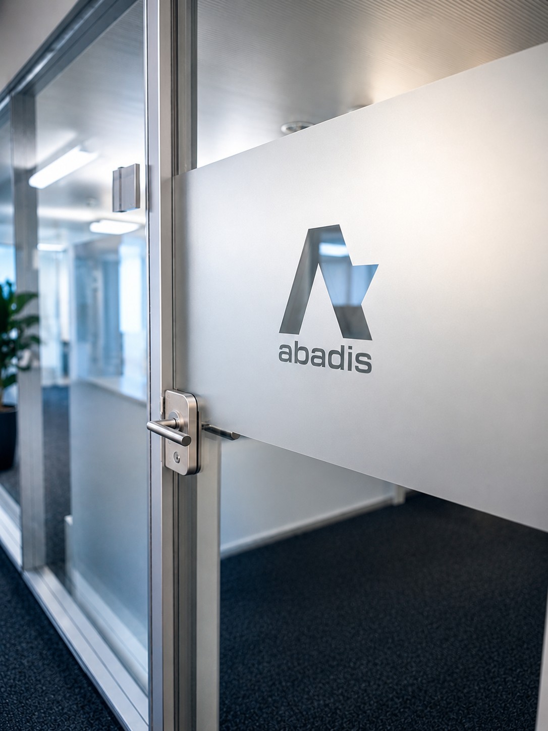 Gläserne Bürotür mit oben mattierter Fläche. Darauf das graue Firmenlogo „abadis“ mit stilisiertem A und Schriftzug. Silberner Griff und Rahmen. Im Hintergrund durch Klarglas helle Büroräume sichtbar.