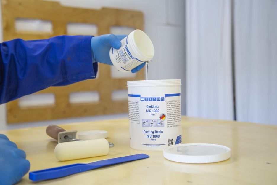 Eine Person in blauen Handschuhen und Arbeitskleidung gießt transparentes WEICON Gießharz MS 1000 aus einem kleineren Behälter in einen großen weißen. Auf dem hellen Holztisch: Farbroller, blauer Spatel und Deckel.