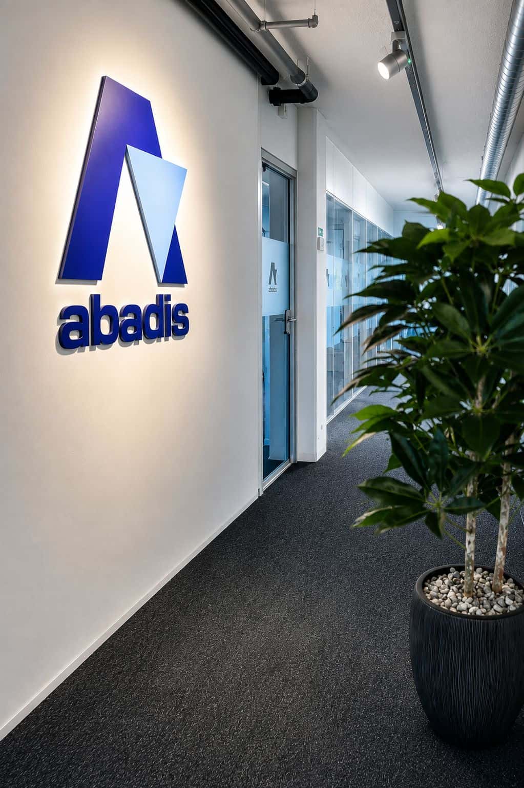 Moderner Bürogang mit beleuchtetem Abadis-Logo links an weißer Wand: blaues stilisiertes „A“ und „abadis“. Rechts vorn große grüne Topfpflanze. Hinten Glastüren zu Büros, Decke mit sichtbaren Leitungen und Strahlern.