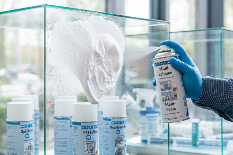 Eine Hand mit blauem Handschuh sprüht den WEICON Multi-Schaum auf eine vertikale Glasfläche. Der dichte, weiße Reinigungsschaum bildet sich und haftet an der Oberfläche. Im unscharfen Hintergrund sind weitere WEICON Spraydosen zu erkennen.