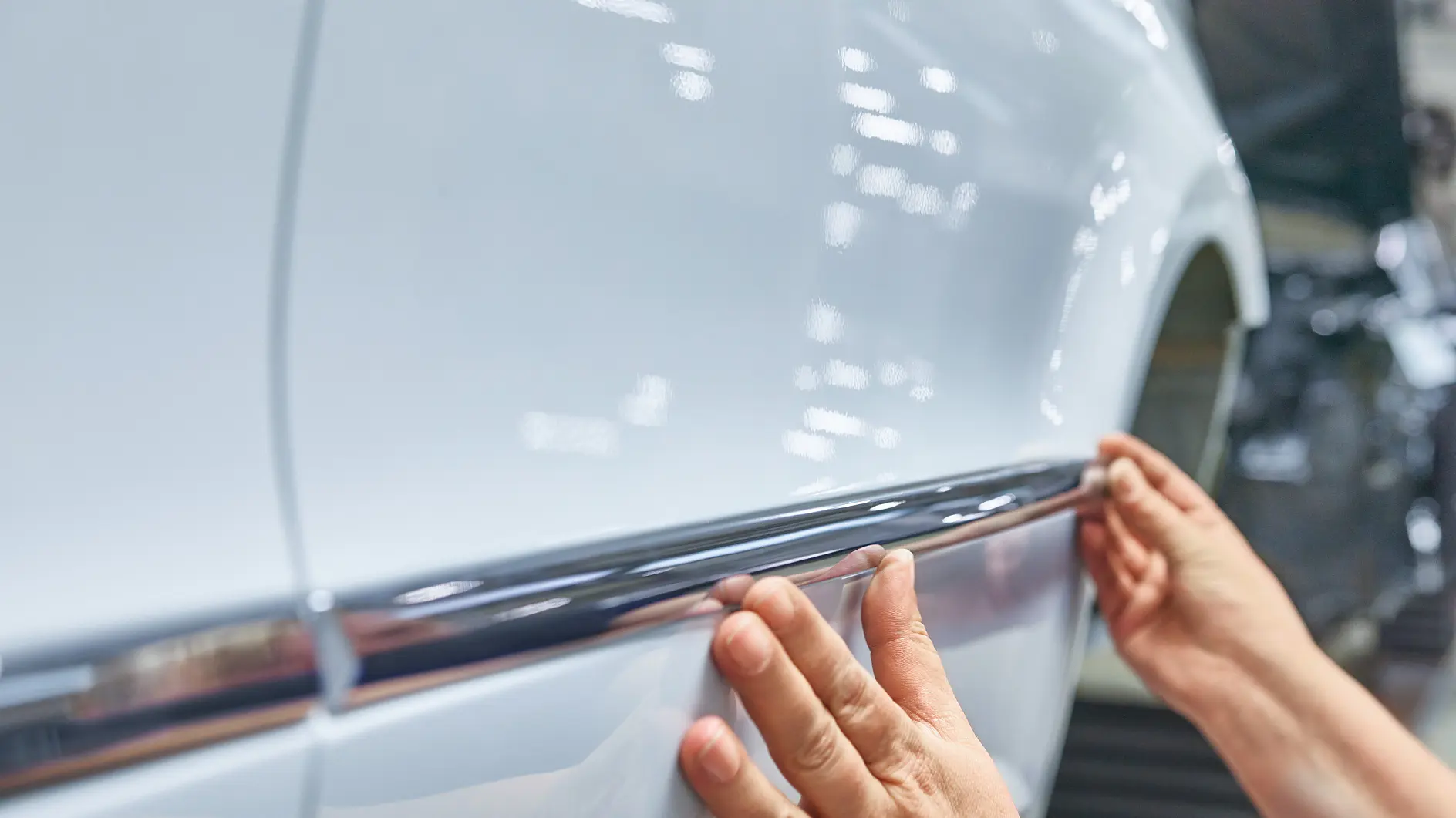 Gros plan : des mains fixent avec précaution une moulure chromée brillante sur le flanc d’une voiture d’un blanc éclatant. La carrosserie reflète des points lumineux, signe d’un travail soigné et précis. Plan d’ensemble : hall de montage.