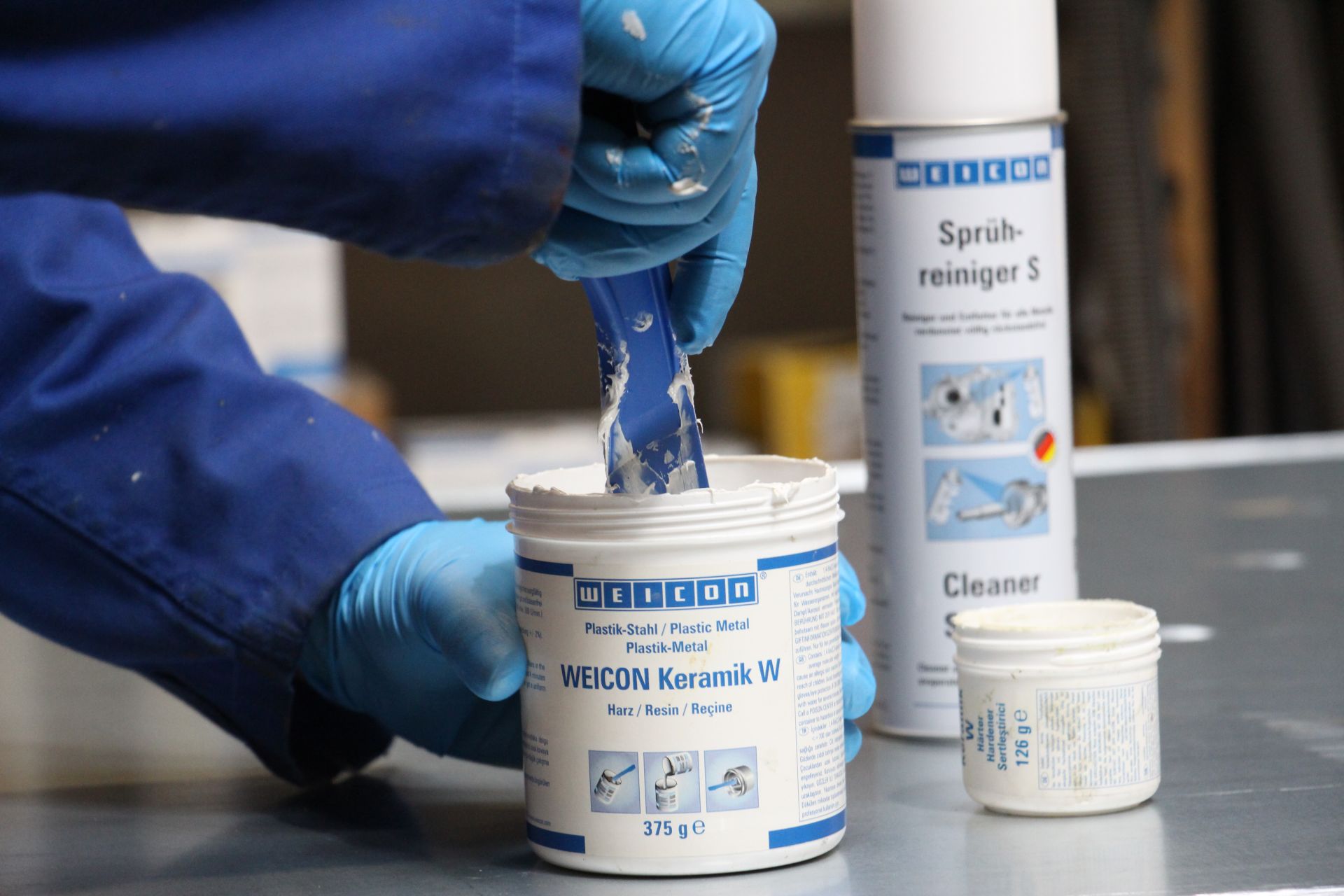Person in blauer Arbeitskleidung und Handschuhen rührt mit blauem Spachtel eine weiße, pastöse Masse aus großem Behälter 'WEICON Keramik W Harz'. Daneben stehen Härter-Behälter und Sprühreiniger auf grauer Metalloberfläche.