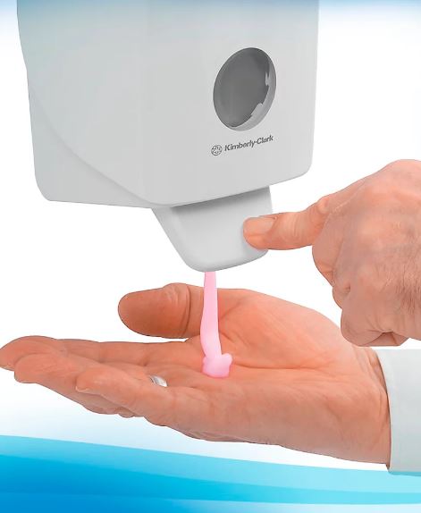 Eine Person drückt einen weißen, wandmontierten Kimberly-Clark-Seifenspender. Rosafarbene Flüssigseife fließt in die offene linke Handfläche, um die Hände zu reinigen. Das Bild betont Handhygiene und regelmäßiges Händewaschen.
