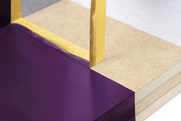 Nahaufnahme einer gemalten Oberfläche mit scharfer Farbkante. Goldfarbenes 3M UV-Abdeckband trennt präzise lebhafte violette Farbe von einer hellbraunen Holzplatte und sorgt für saubere Farblinien; ein senkrechter Streifen betont die Exaktheit.