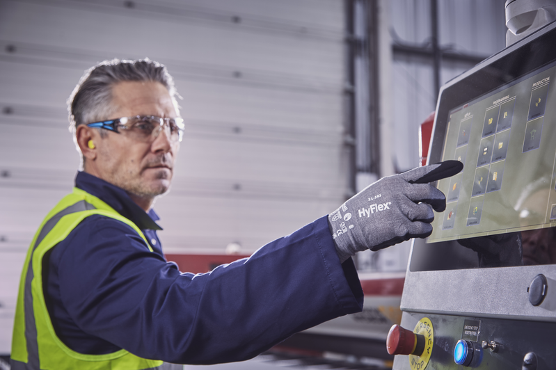 Ein Arbeiter mit Schutzbrille, Gehörschutz und Warnweste bedient in einer Fabrikhalle den Touchscreen einer Maschine. Er trägt graue HyFlex Schnittschutzhandschuhe mit Touchscreen-Fähigkeit und wählt eine Option aus.