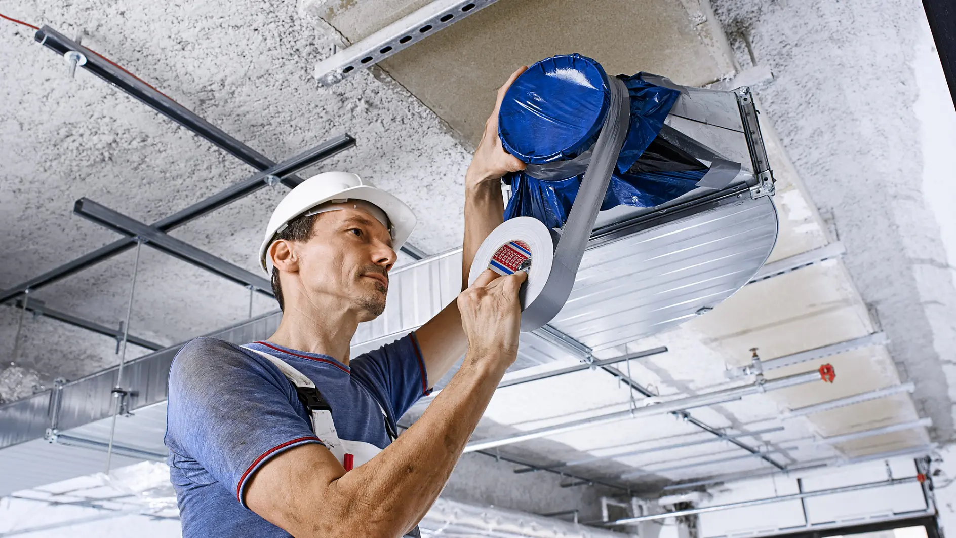 Un ouvrier du bâtiment, coiffé d'un casque de sécurité, fixe un conduit de ventilation au plafond. Il utilise du ruban adhésif gris pour calfeutrer un élément recouvert d'un film bleu. Chantier avec un plafond brut et des rails métalliques.