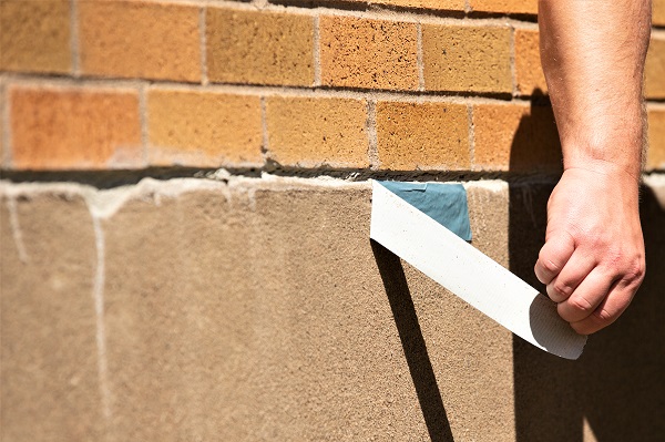 Eine Hand bringt ein blau-graues 3M Hochleistungs-Gewebeklebeband zwischen eine braune Ziegelwand und eine darunter liegende beige Betonfläche an. Ein weißer Schutzstreifen wird vom 3M Performance Plus 8979 abgezogen.