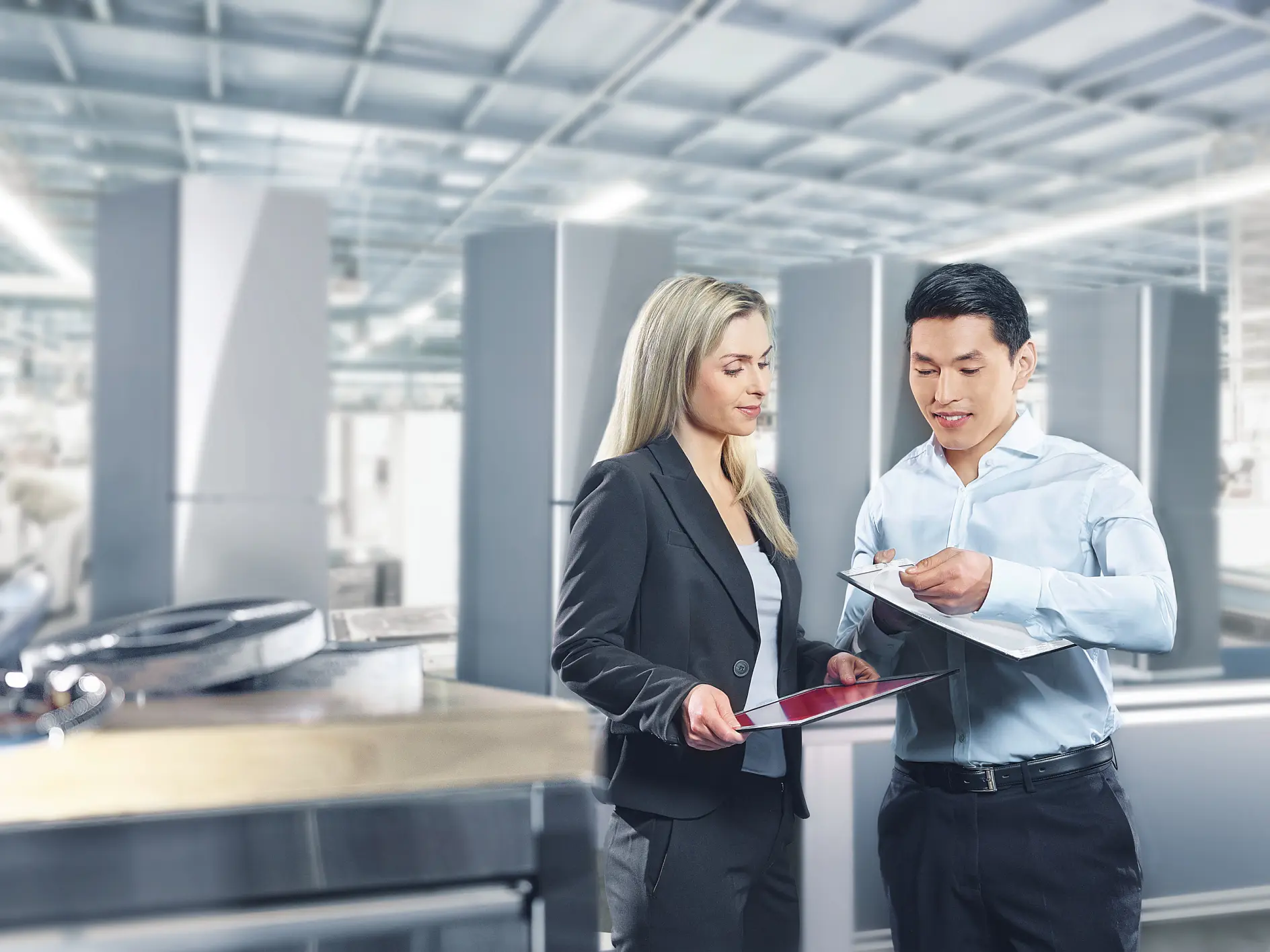 Deux professionnels côte à côte dans un hall industriel moderne : une femme blonde en tailleur sombre et un homme aux cheveux foncés en chemise bleu clair. Elle examine des documents ; lui tient un bloc-notes, tandis qu'elle porte un dossier rouge.