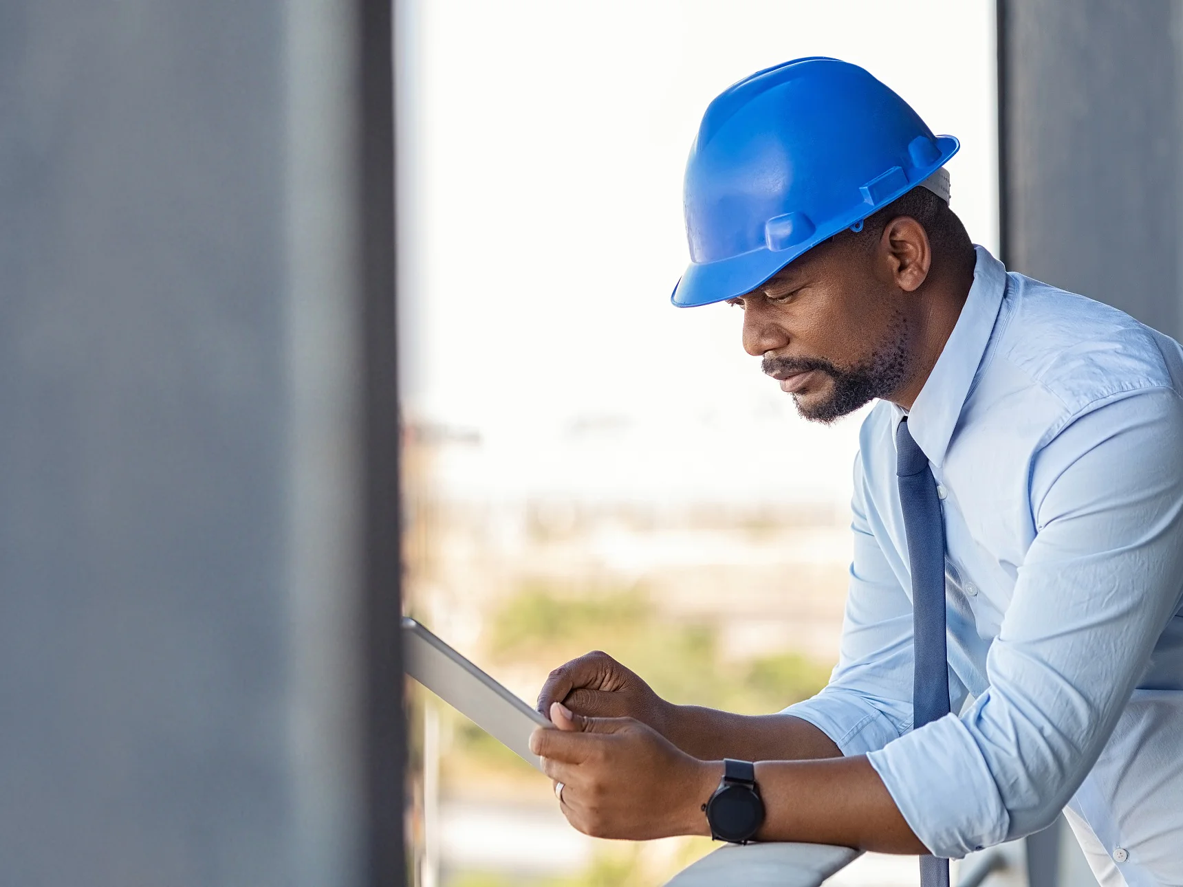 Un homme à la peau foncée, casque de chantier bleu, chemise bleu clair et cravate bleu foncé, se penche sur un balcon. Il regarde une tablette tenue à deux mains et porte une smartwatch noire au poignet gauche. Fond urbain flou.